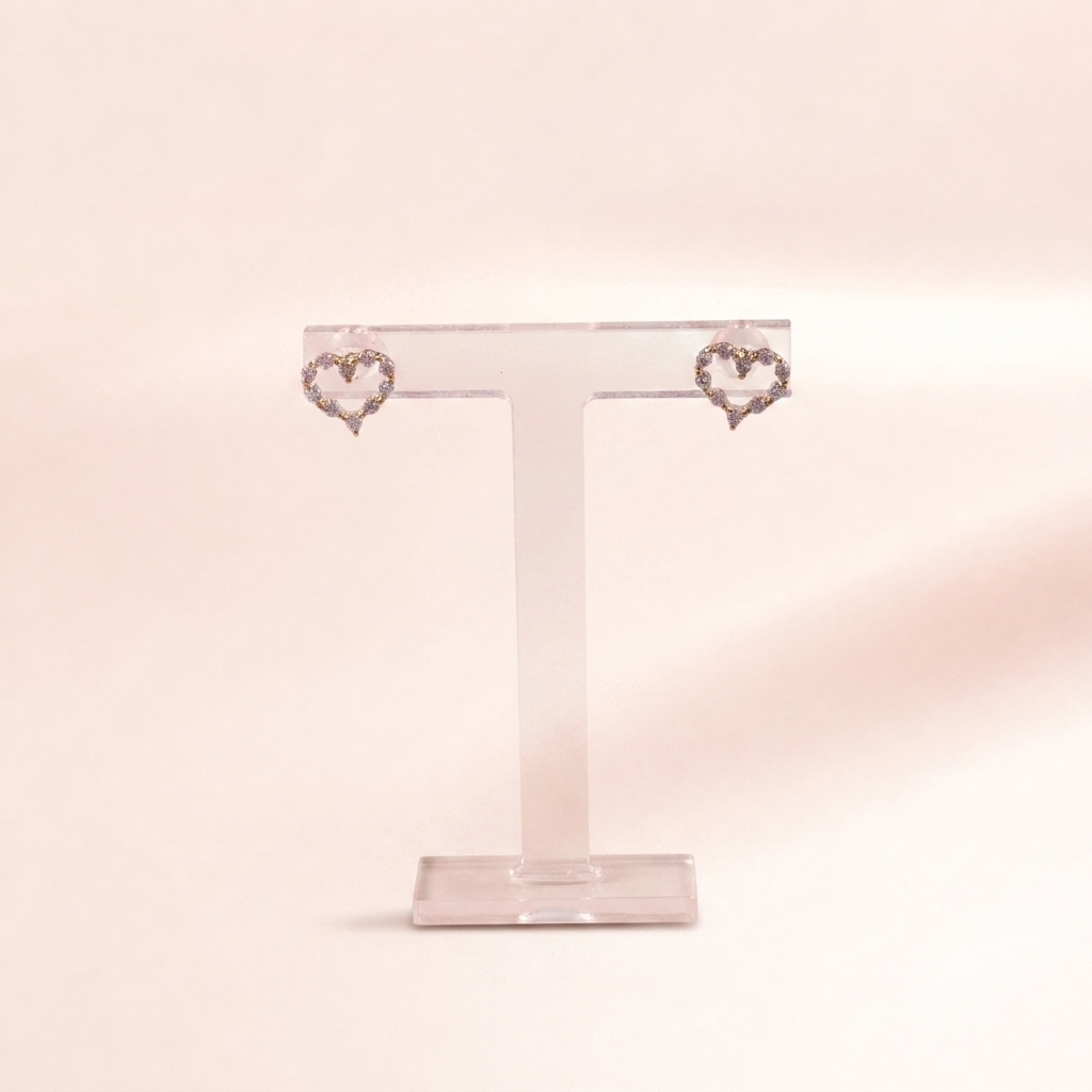 A pair of silver heart-shaped stud earrings with small clear stones, displayed on a clear acrylic T-stand against a soft, pale pink background.