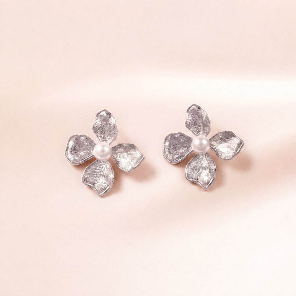 A pair of silver flower-shaped stud earrings with textured petals and a small white pearl in the center, displayed on a soft, light pink background.
