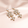 A pair of gold-tone dangle earrings with clear marquise and round crystals, arranged on a soft blush pink satin fabric background.
