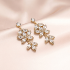 A pair of gold-tone dangle earrings with clear marquise and round crystals, arranged on a soft blush pink satin fabric background.