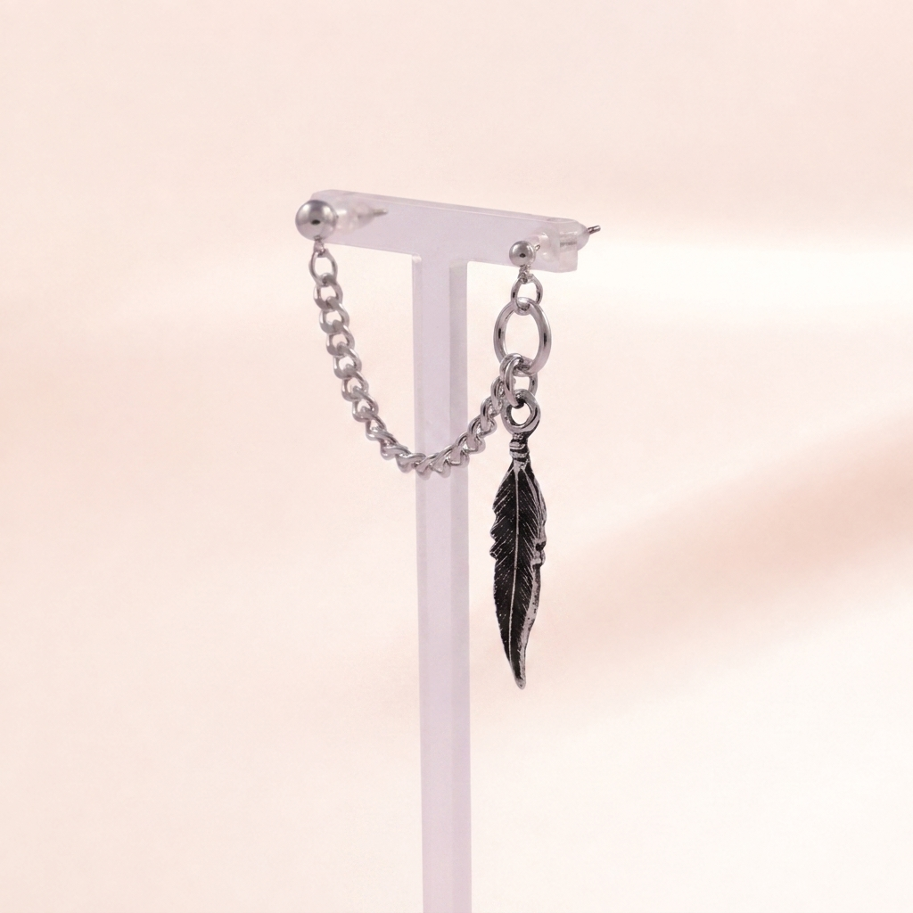 A silver-toned dangle earring with a small chain and a feather-shaped charm, displayed on a clear jewelry stand against a soft beige background.