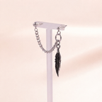 A silver-toned dangle earring with a small chain and a feather-shaped charm, displayed on a clear jewelry stand against a soft beige background.
