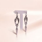 A pair of silver-toned dangle earrings displayed on a clear T-stand against a soft warm beige background. Each earring features a circular ring with textured spiral details, a short chain, and a pointed cone-shaped drop.