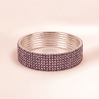 A wide silver-toned bangle bracelet embellished with multiple rows of small round purple rhinestones, shown on a soft beige background.