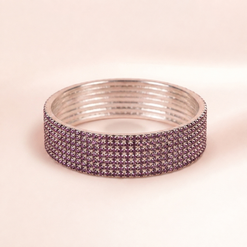 A wide silver-toned bangle bracelet embellished with multiple rows of small round purple rhinestones, shown on a soft beige background.