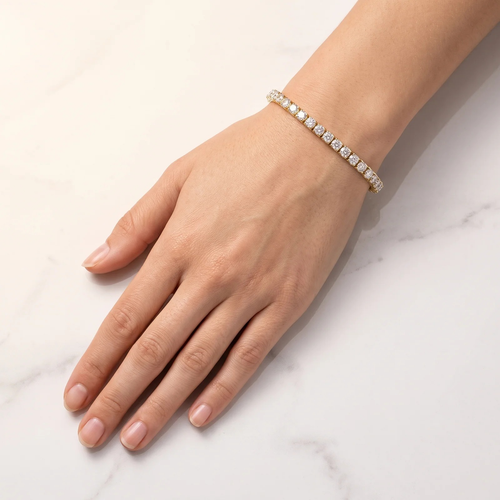 A close-up of a person's hand and wrist wearing a delicate gold tennis bracelet with clear square-cut stones, set against a light marble background.