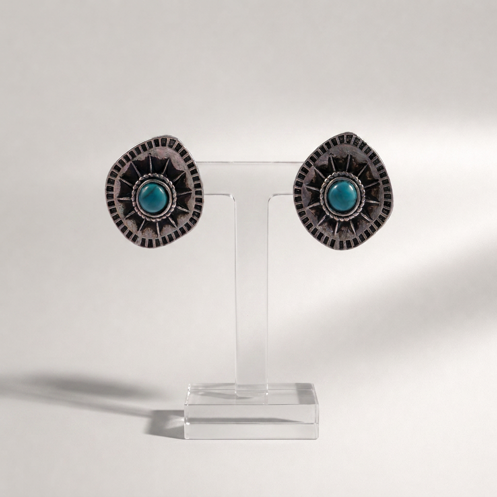 A pair of silver-toned statement earrings with oval turquoise centers and intricate sunburst detailing, displayed on a clear acrylic stand against a light gray background.