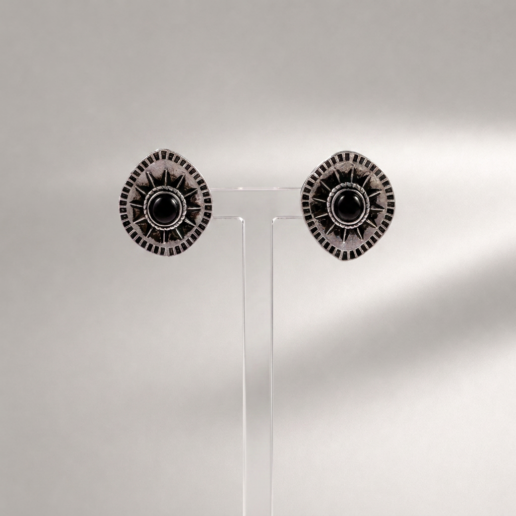 A pair of round silver-toned earrings with black circular centers and radiating textured details, displayed on a clear acrylic stand against a light gray background.