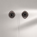 A pair of round silver-toned earrings with black circular centers and radiating textured details, displayed on a clear acrylic stand against a light gray background.
