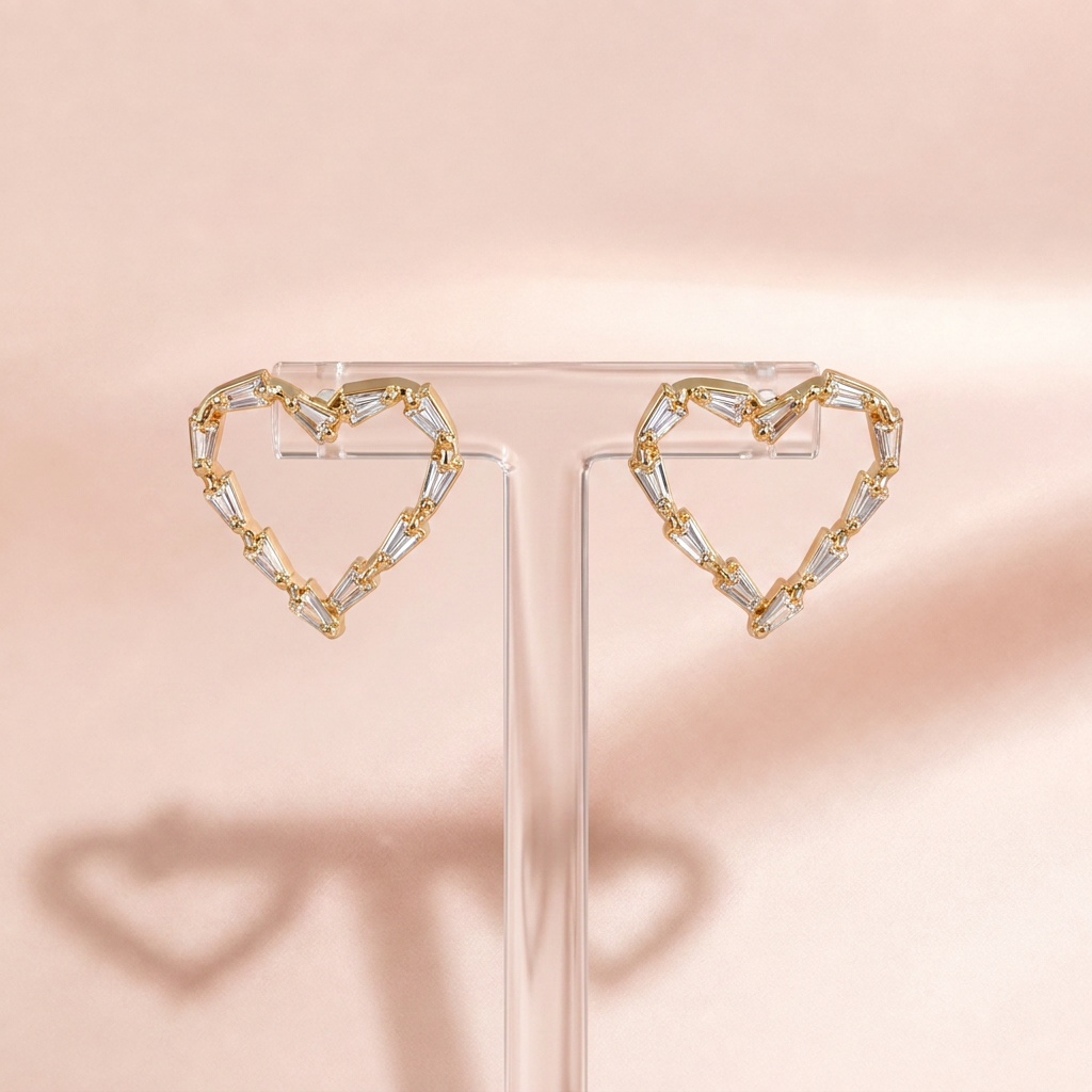 A pair of gold heart-shaped stud earrings with clear faceted baguette and round crystals, displayed on a clear acrylic stand against a soft pink background.