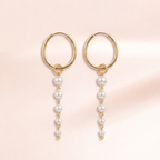A pair of gold hoop earrings with dangling pearl bead chains, displayed on a soft pink background.