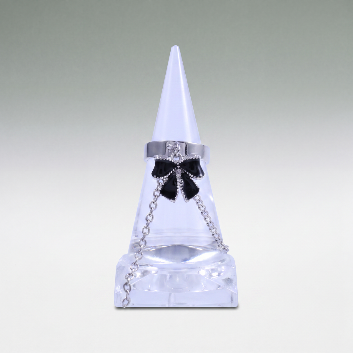 A silver-toned ring with a black bow charm and clear rhinestone accents, displayed on a clear cone jewelry stand against a light gray gradient background.