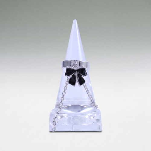 A silver-toned ring with a black bow charm and clear rhinestone accents, displayed on a clear cone jewelry stand against a light gray gradient background.